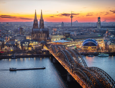 A breathtaking view of the Cologne skyline right after sunset featuring the iconic Cologne Cathedral and the city's famous bridge. The warm hues of the evening sky create a stunning scene in Germany.